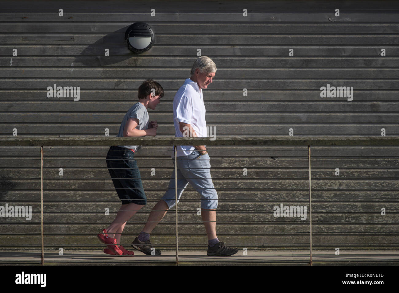 A man on a walkway walking into the wind with his son sheltering behibd him. Stock Photo