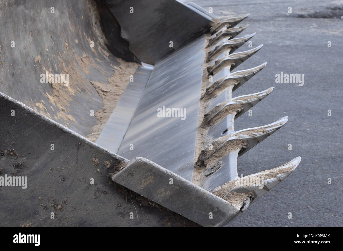 Empty bucket of excavator with traces of sand and soil on the glossy teeth and surfaces against asphalt background. Stock Photo
