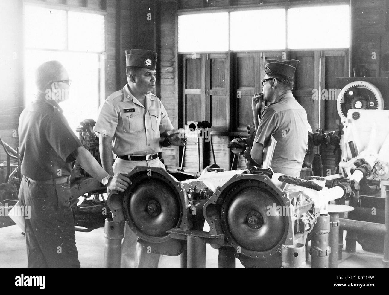 African-american lieutenant standing with two other American servicemen during the Vietnam War, in a heavy vehicle machine shop, one man smoking a cigarette, the other man resting his hand on a part for a truck, the lieutenant gesturing and in mid speech, 1969. Stock Photo
