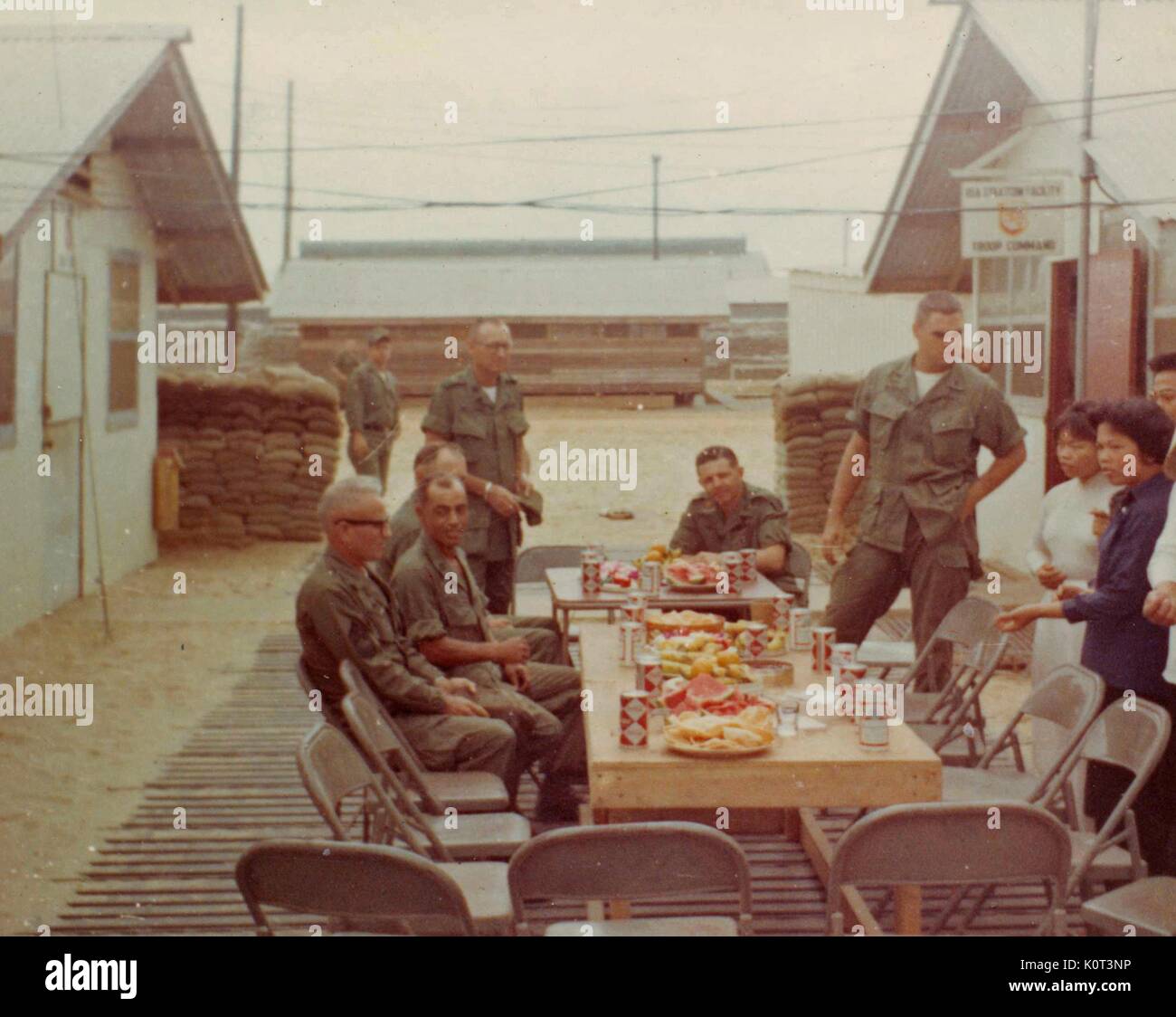 Tet Holiday, American soldiers enjoying a meal at a long table to celebrate the holiday at their base in Vietnam during the Vietnam War, drinks and other food items visible on the table, several South Vietnamese woman standing nearby, 1965. Stock Photo