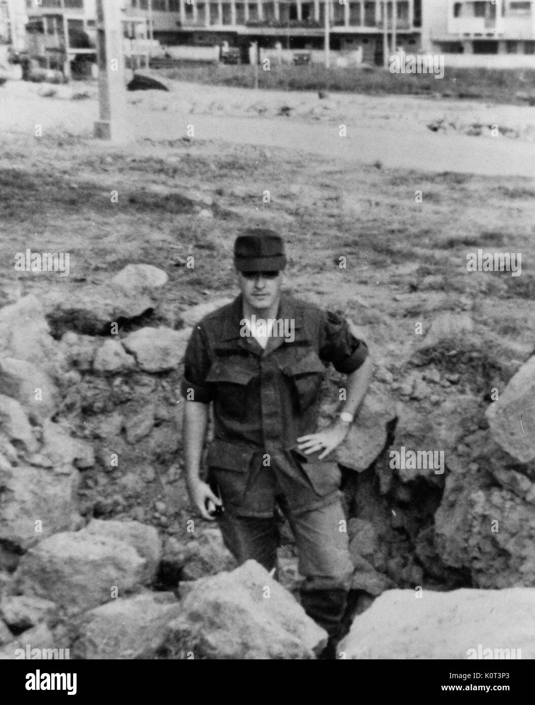 American Marine standing in a fox hole, wearing military uniform, with a cap obscuring his face, one hand on his hip, near a dirt road in Vietnam during the Vietnam War, 1965. Stock Photo