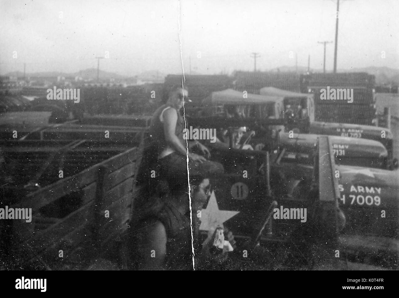 A photograph of two United States Army servicemen sitting in the bed of an M35 military truck, other trucks, building materials, and the Army base they are located on are seen in the background, Vietnam, 1967. Stock Photo