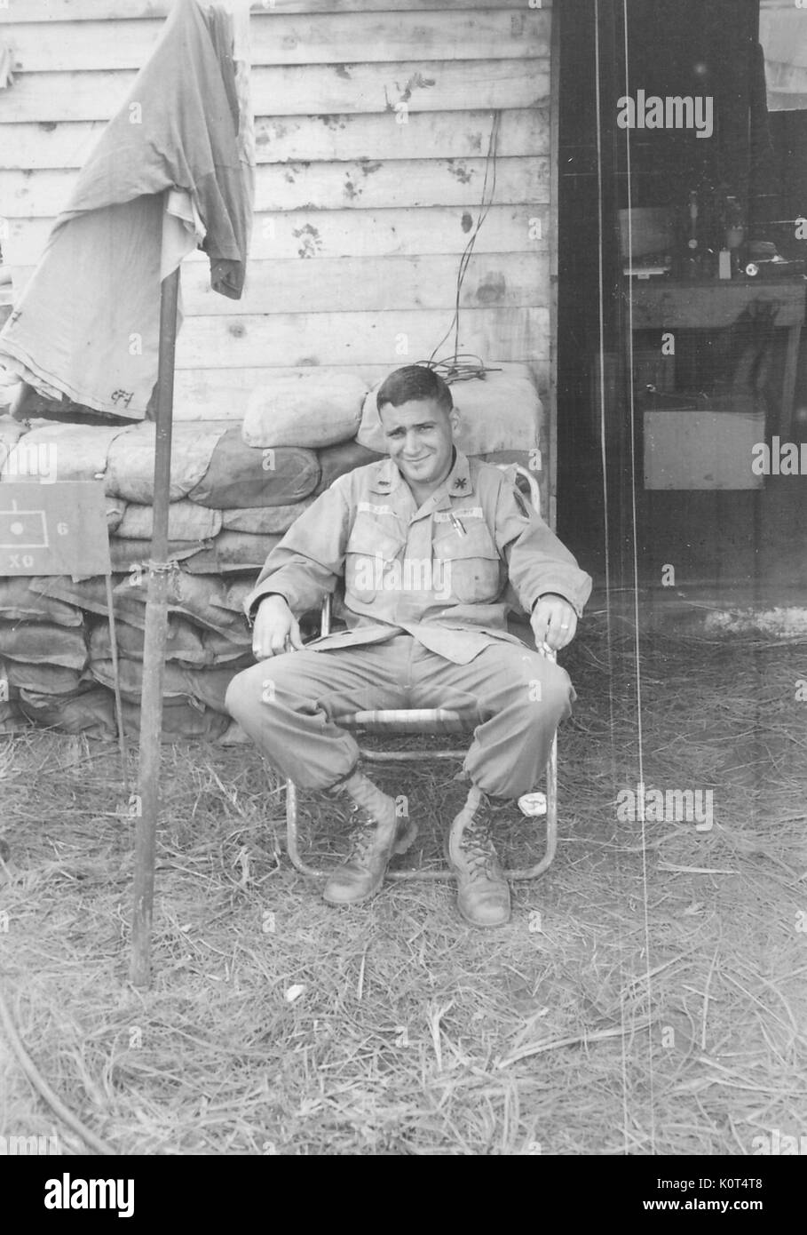 A United States Army serviceman relaxing in a lawn chair, he is smoking a cigarette near the entrance to a wooden building where equipment is visible spread out on a table, Thailand, 1967. Stock Photo