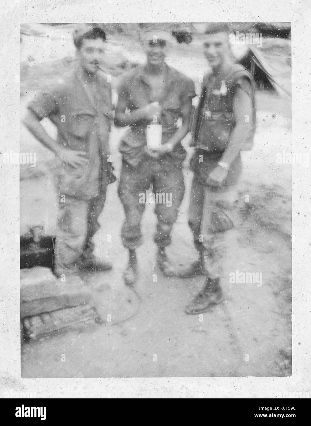 An out-of-focus photograph of three members of the United States Marine Corps standing together and smiling, the soldier in the middle is holding an large glass bottle, a tent and a truck can be seen in the background, Vietnam, 1967. Stock Photo