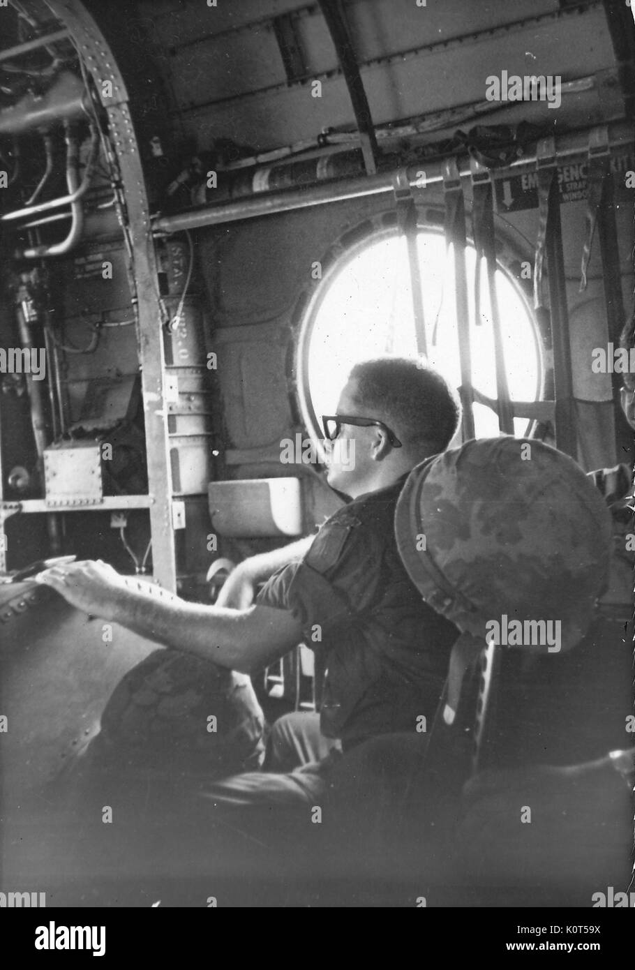 A United States Army serviceman seated near a large window on an airplane, he has his gear between his legs and a helmet hanging from his chair, Vietnam, 1967. Stock Photo