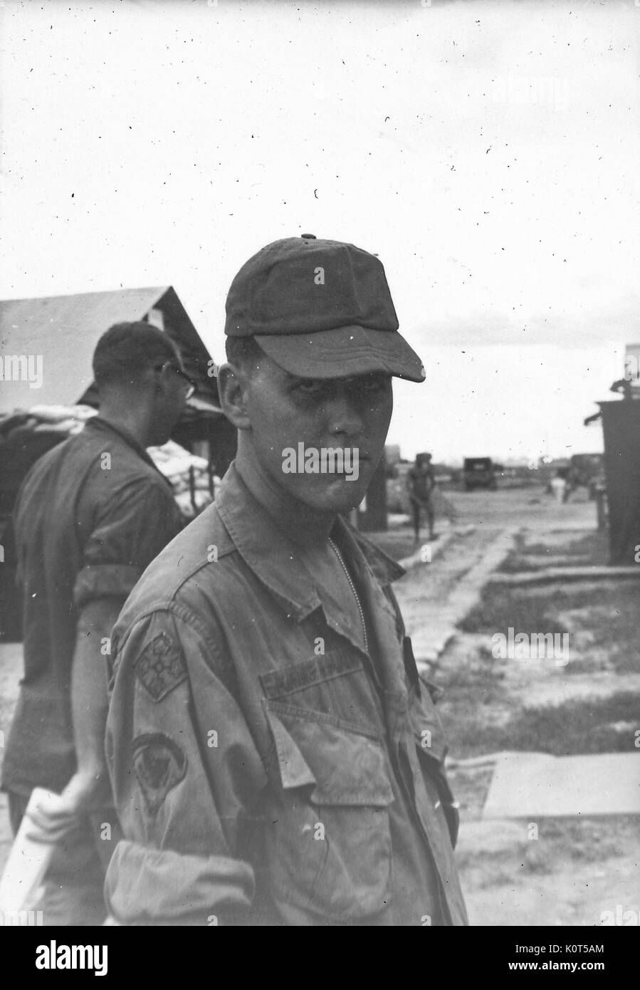 A soldier putting on a serious face while posing in his uniform, other soldiers can be seen in the background on the military base, Vietnam, 1967. Stock Photo