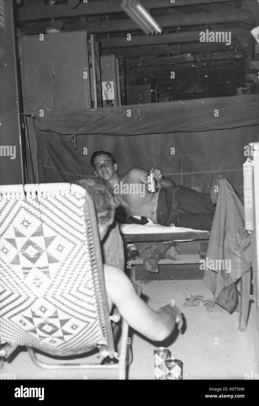 A photograph of two United States Army serviceman socializing in their barracks, one soldier is lying in bed and holding a can of Coca Cola, another serviceman sits in a lawn chair and has several Coke cans on the ground beside him, Vietnam, 1967. Stock Photo
