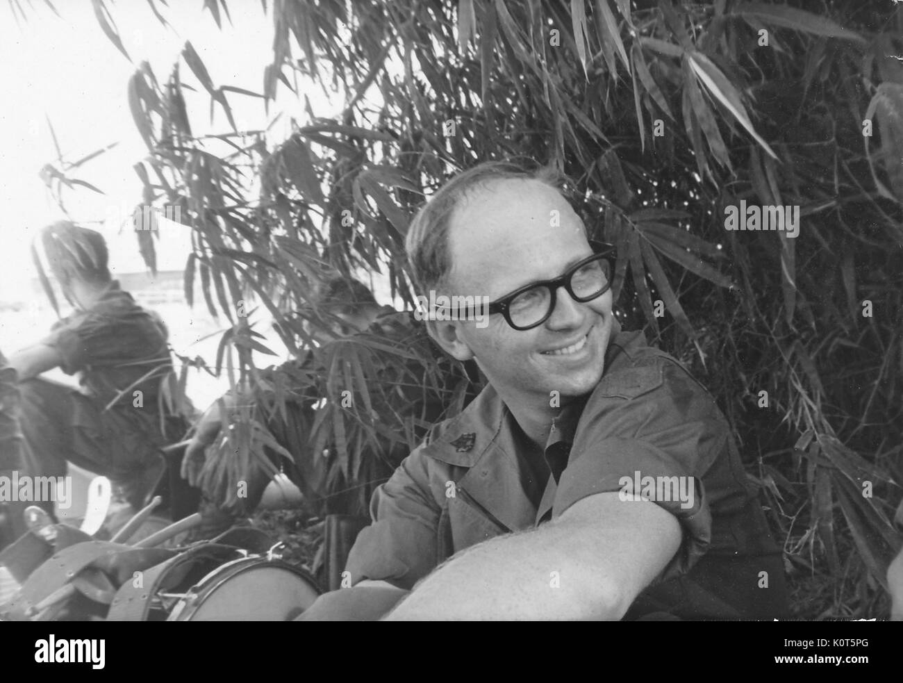 A member of the United States Army First Infantry Division sitting in the shade created by some tall tropical foliage, drumming equipment and another soldier resting in the shade are seen nearby, Vietnam, 1967. Stock Photo