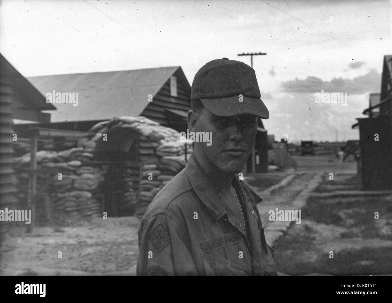 A photograph a United States Army serviceman wearing his jungle jacket, cap and a stoic expression, wooden buildings and a sandbag reinforced structure are visible behind him on the base, Vietnam, 1967. Stock Photo