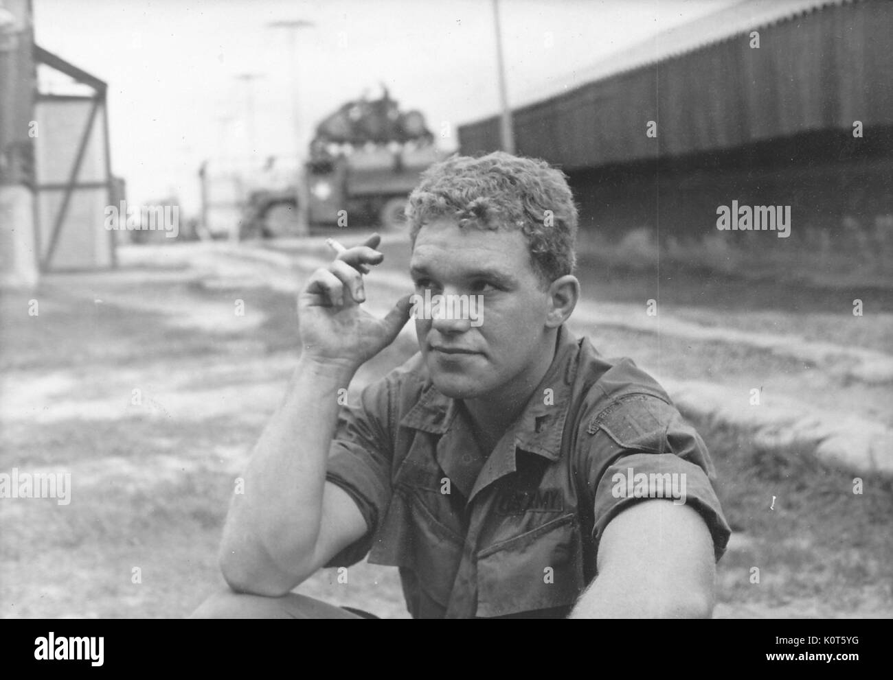 A United States Army serviceman sitting on the ground and smoking a cigarette, buildings and utility poles can be seen on the base behind him, Vietnam, 1967. Stock Photo