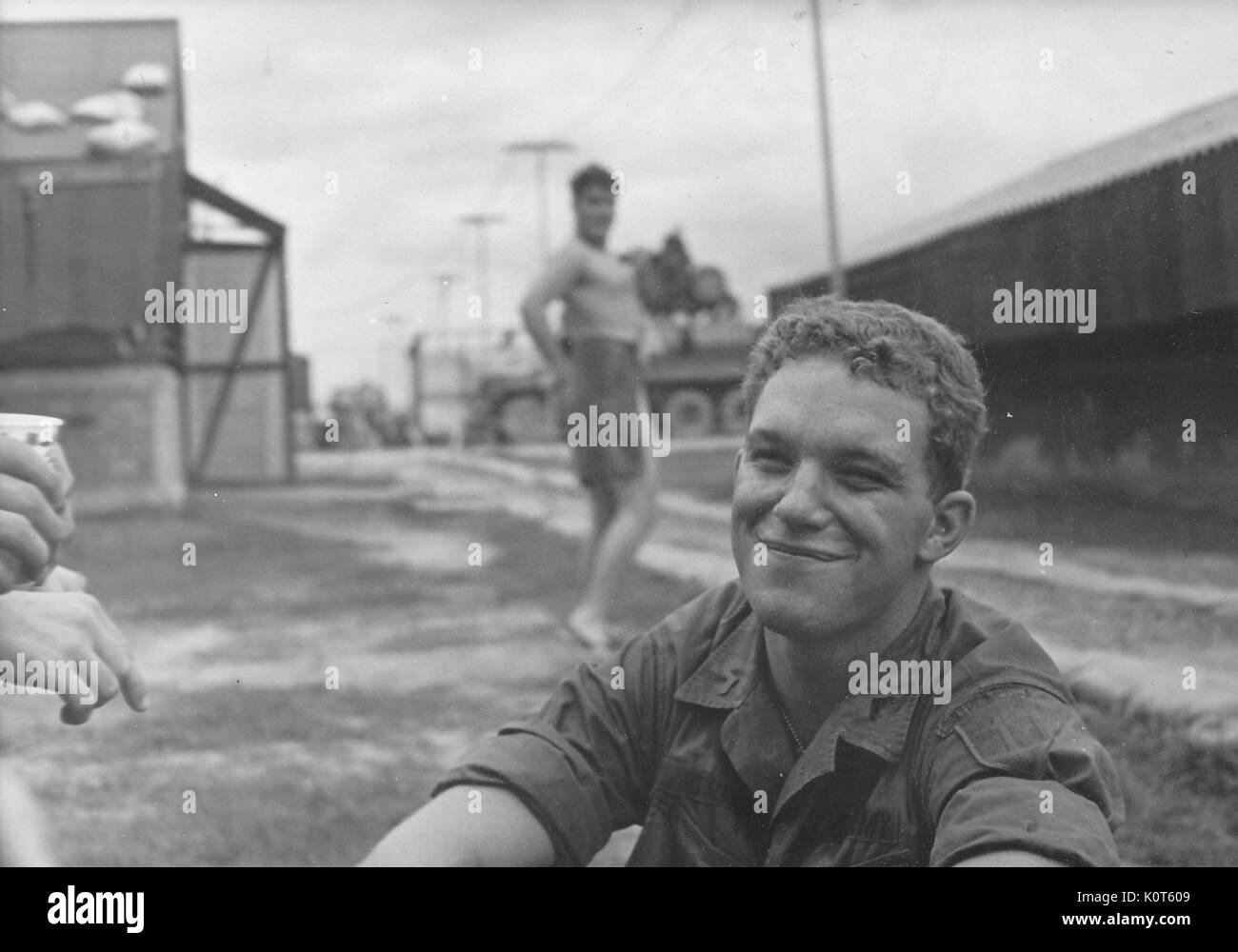 A United States Army soldier smiling while he is sitting on the ground, another soldier wearing only a towel in the background is trying to get the attention of the photographer by striking the pose of a pin-up girl, Vietnam, 1967. Stock Photo