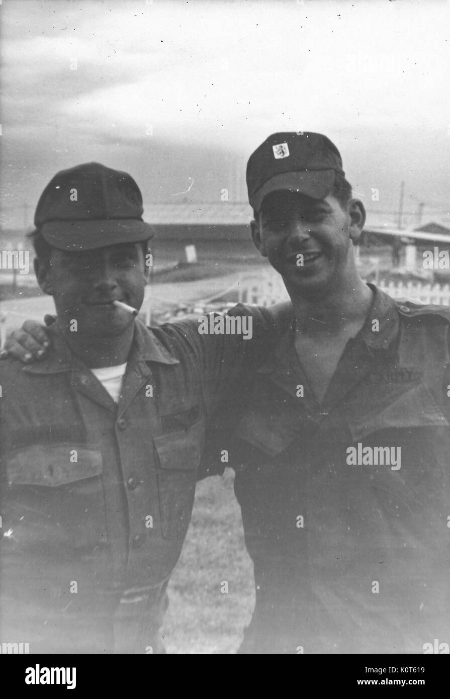A photograph of two United States Army soldiers posing with their arms around each others shoulders, the serviceman on the left is smoking a cigarette, a portion of the base case be seen in the background, Vietnam, 1967. Stock Photo