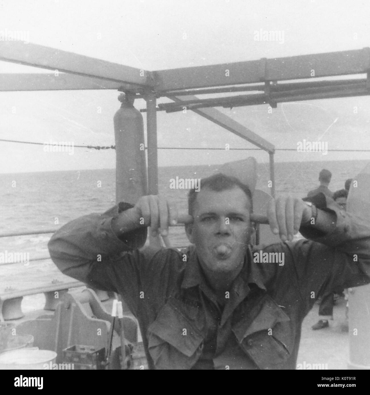 A United States soldier sticks his tongue out and makes a funny face while posing aboard a large ship, Vietnam, 1966. Stock Photo