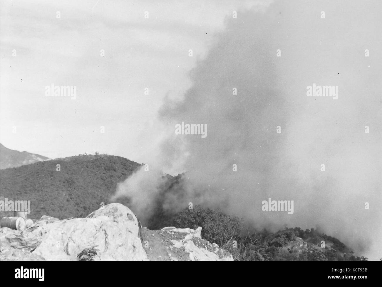 In the jungles of Vietnam, a plume of smoke billows out from a mountainside near several oil drums, 1966. Stock Photo