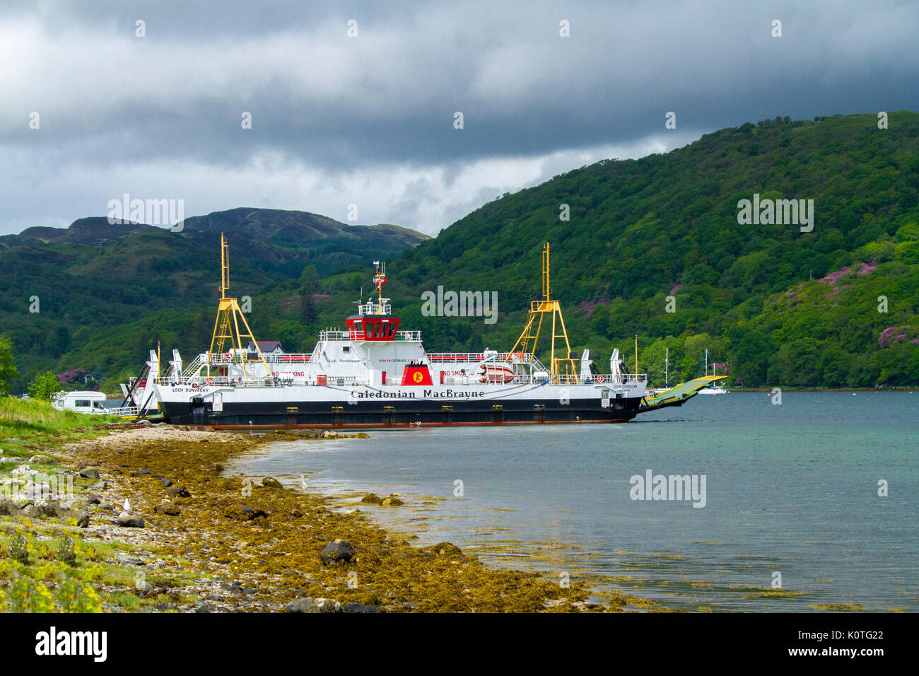 Vehicular ferry, Caledonia MacBrayne, moored at island of Bute with wooded mountains of mainland of Scotland beside calm blue waters of ocean strait Stock Photo
