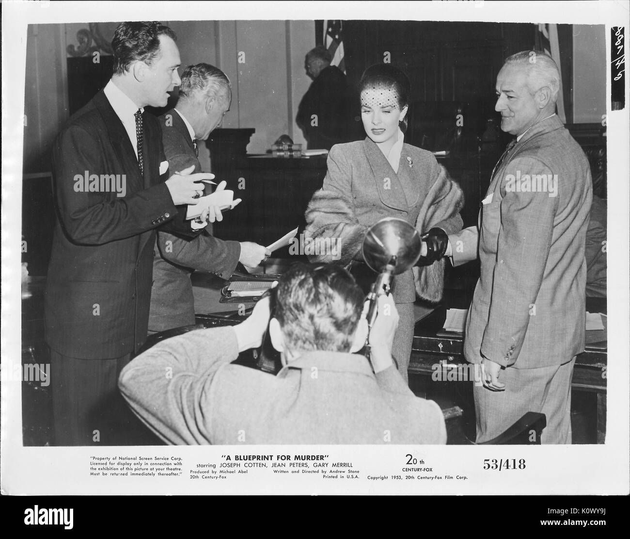 A movie still scene from 'A Blueprint For Murder' (1953 20th Century Fox thriller film), showing an elegant woman being interviewed and photographed by some press people while she hold on to a man with white hair for moral support, 1953. Stock Photo
