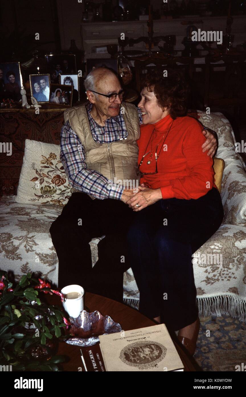 Mature couple smiling and embracing on a couch, holding hands and looking into each others eyes, in a cluttered suburban home with photos of their family visible in the background, the woman wearing an orange shirt, the man with a plaid shirt and vest, 1960. Photo credit Smith Collection/Gado/Getty Images. Stock Photo