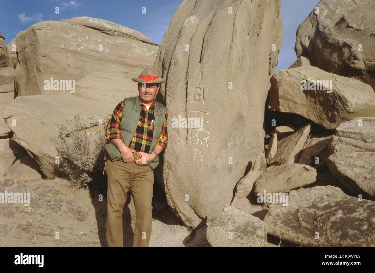 Hunter wearing cowboy hat, flannel vest and overalls standing and leaning against a rock on which he has carved his initials and the year, 1974. Stock Photo