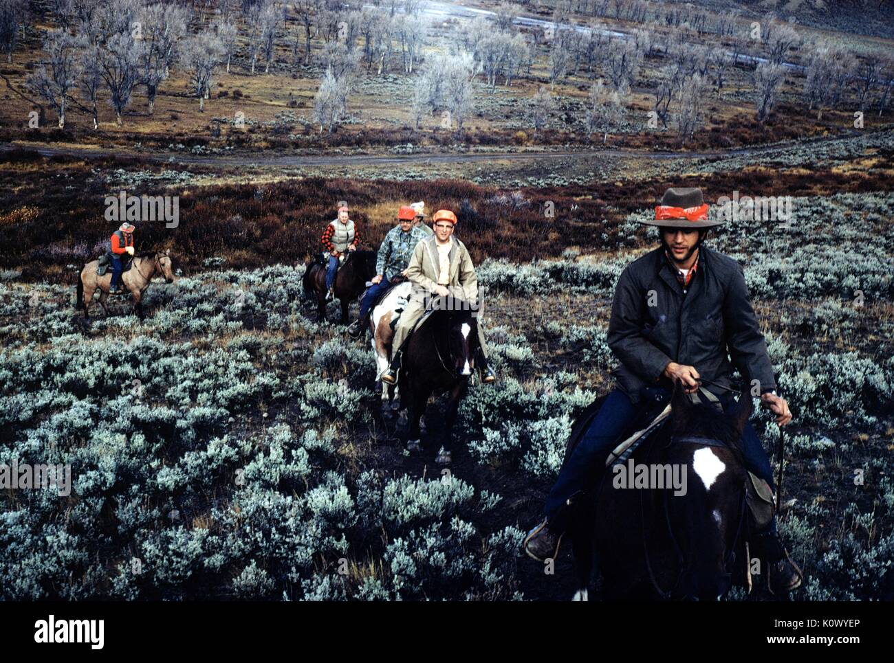 Group of hunters on horseback, riding through scrubgrass in a line during a hunting trip, wearing cowboy hat and smiling, 1971. Stock Photo
