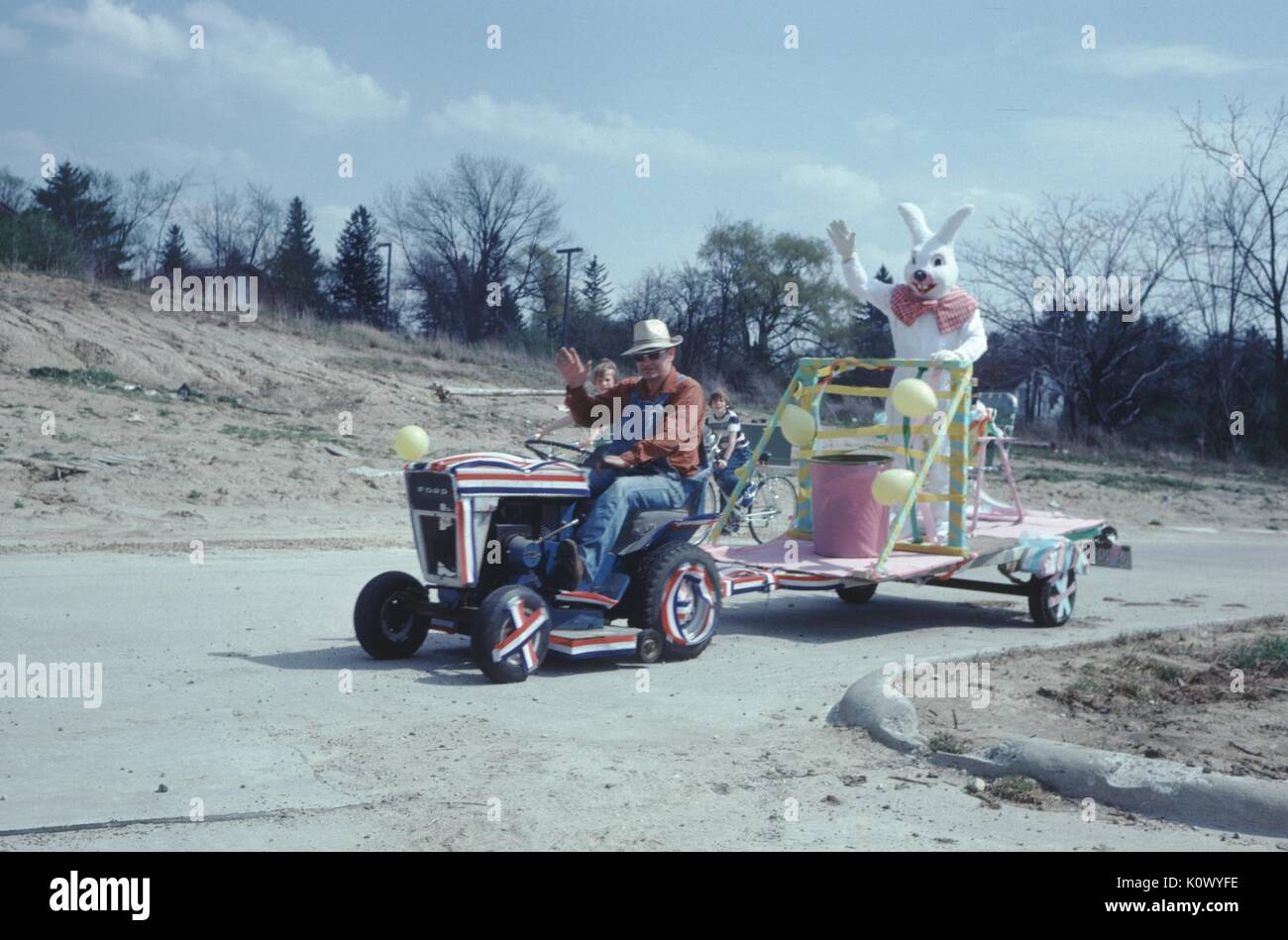 Parade float, a man in a cowboy hat sitting on a riding lawnmower and waving, wearing a cowboy hat, pulling a parade float on which another man stands, wearing a white rabbit costume and waving, during a July 4th parade, paper bunting on the tractor, 1971. Photo credit Smith Collection/Gado/Getty Images. Stock Photo