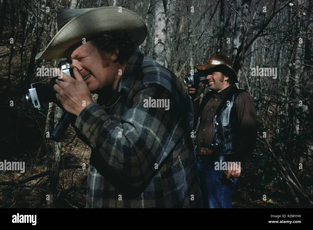 Two hunters in the woods, dressed in flannel and cowboy hats, one looking through the lens of a 35mm still camera, the other looking through the lens of an 8mm home movie film camera, surrounded by trees, Parched Corn Creek, Kentucky, 1975. Photo credit Smith Collection/Gado/Getty Images. Stock Photo