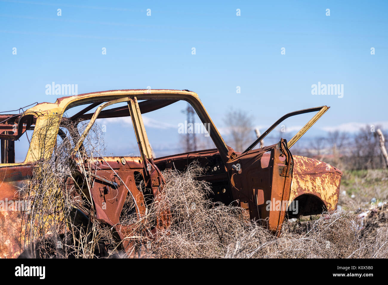 Abandoned and rusty wreckage of an yellow vintage Soviet Russian car in the middle of dry hay with scenic ice top mountains in Southern Armenia. Stock Photo