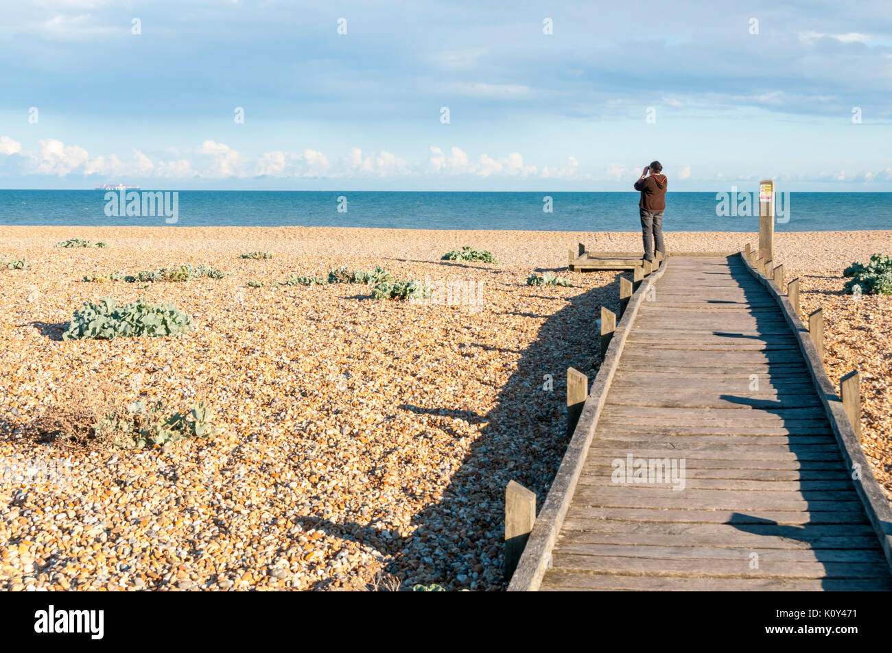 A woman looking out to sea with binoculars from the end of a boardwalk across the shingle beach at Dungeness Stock Photo