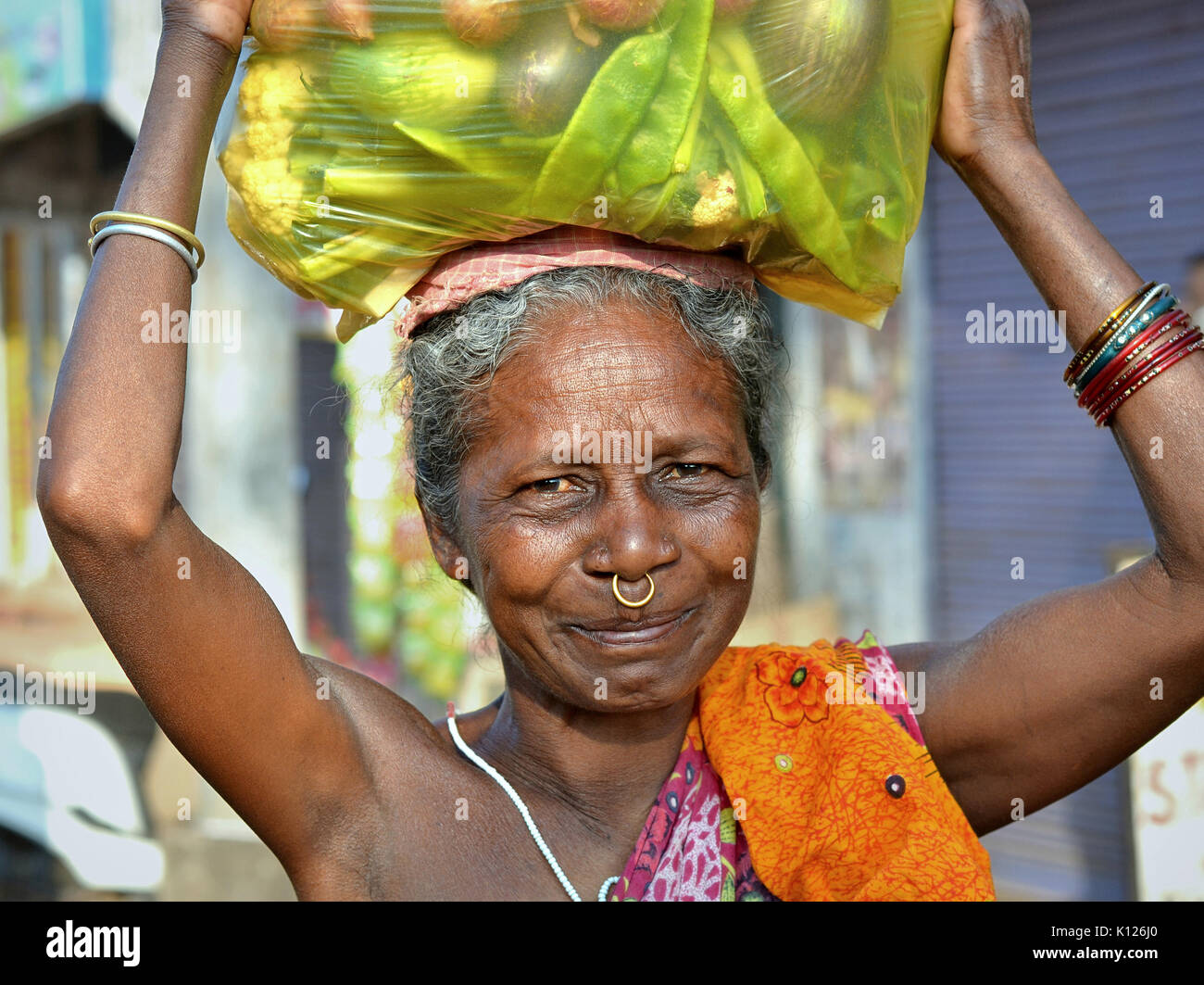 Elderly Indian Adivasi woman (Orissan tribal woman) with one golden nose ring carries on her head a heavy plastic bag full of assorted vegetables. Stock Photo
