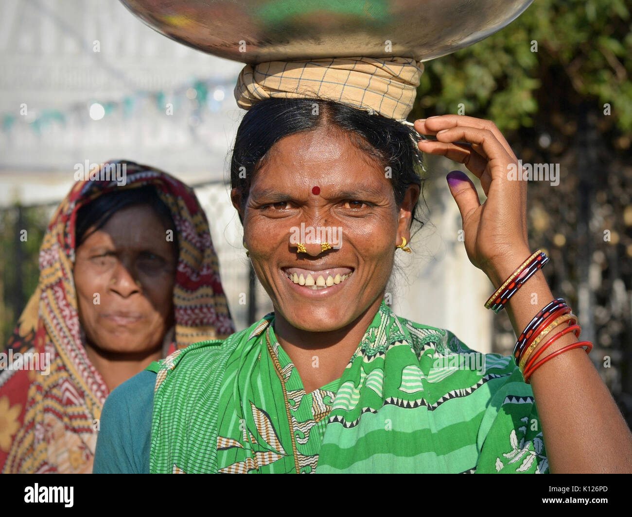 Indian Adivasi woman (Orissan tribal woman) with two distinctive golden nose studs balances on her head a heavy metal bowl and smiles for the camera. Stock Photo