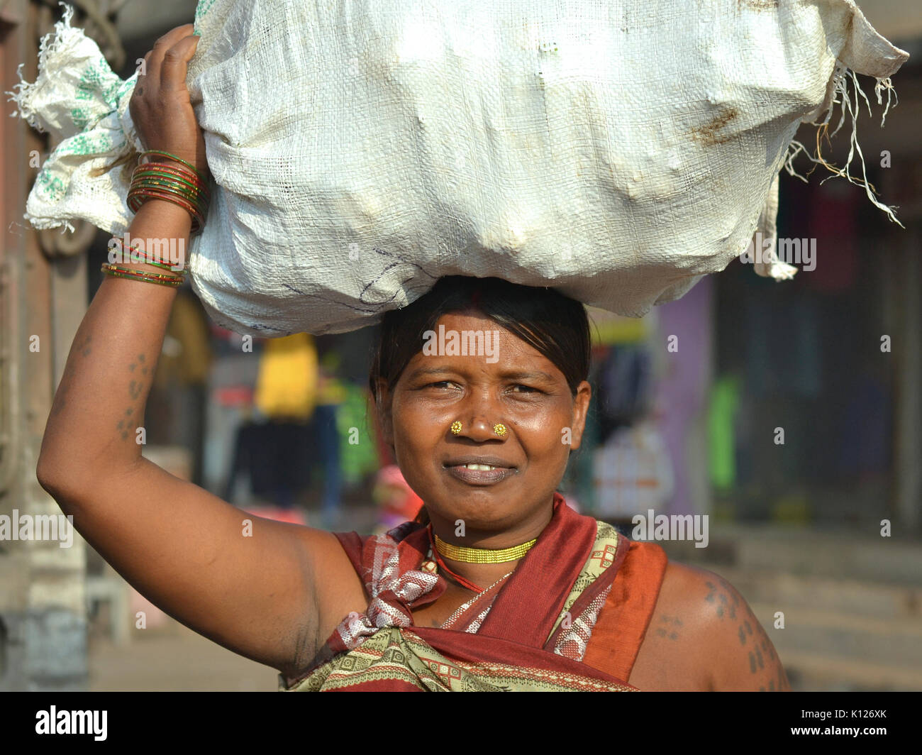 Indian Adivasi woman (tribal woman) with two distinctive golden nose studs balances on her head a heavy bag of vegetables and smiles for the camera. Stock Photo
