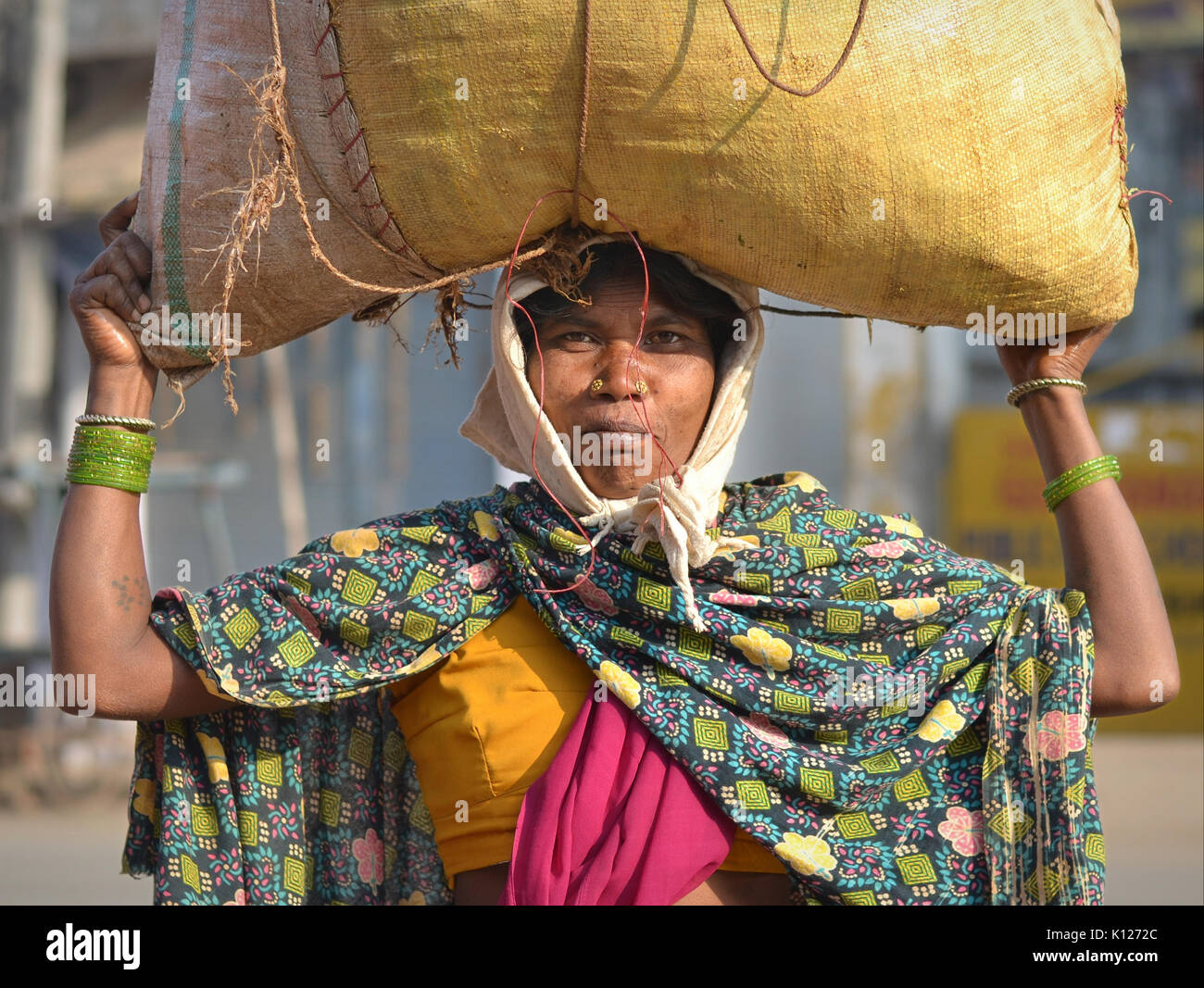 Indian Adivasi woman (tribal woman) with two distinctive golden nose studs carries on her head a bag of vegetables and poses for the camera. Stock Photo