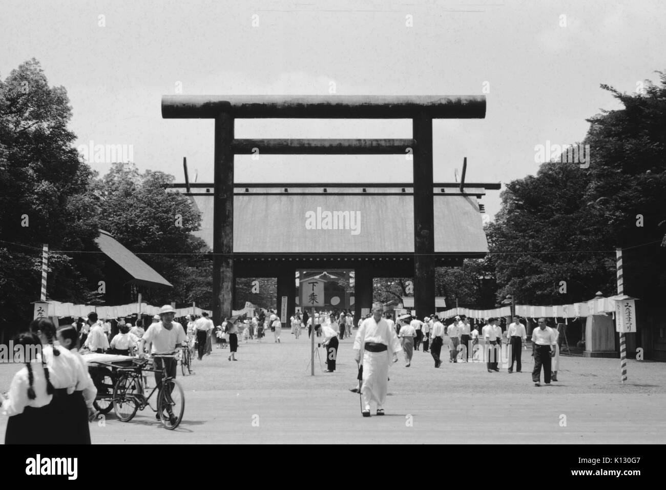 Crowd of Japanese visitors walking on a street in front of the gateway arch of a temple, wearing black and white attire, one man pushing a bicycle, another holding a wooden sign, unfurled prayer scrolls visible, Japan, 1952. Stock Photo