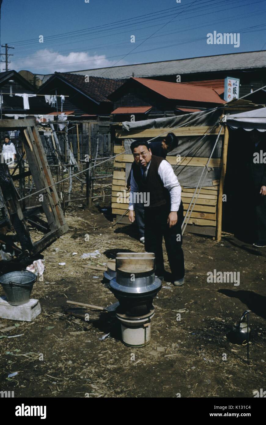 Japanese man in black vest and white shirt, bending over and heating a vessel on an outdoor heater, a teapot visible on the ground, Japan, 1952. Stock Photo
