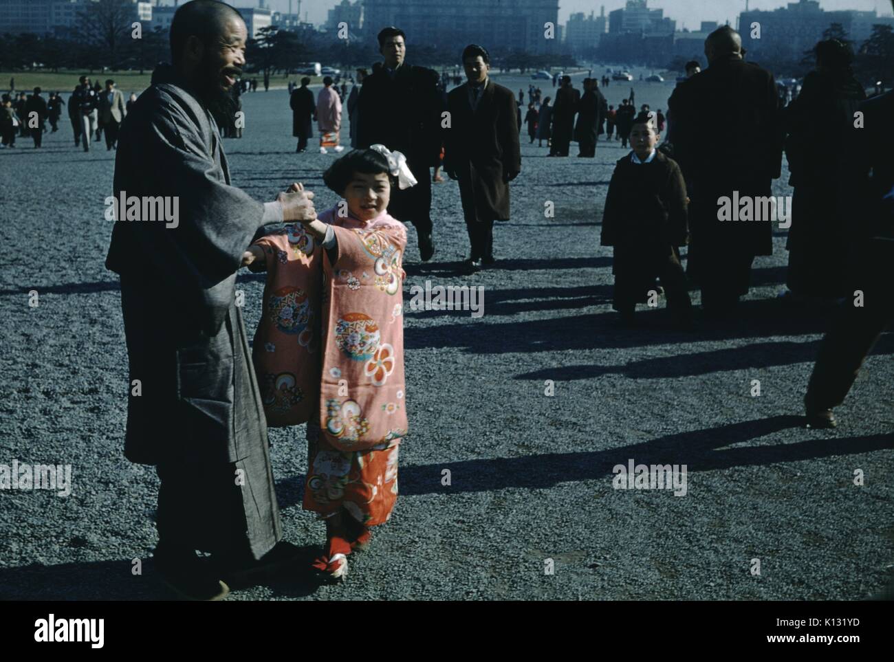Japanese man with bald head, robe and long beard smiling and holding the hands of his young daughter, who wears a traditional pink Kimono and Geta sandals, in a public square, with a large number of people walking past in groups of two to three, 1952. Stock Photo