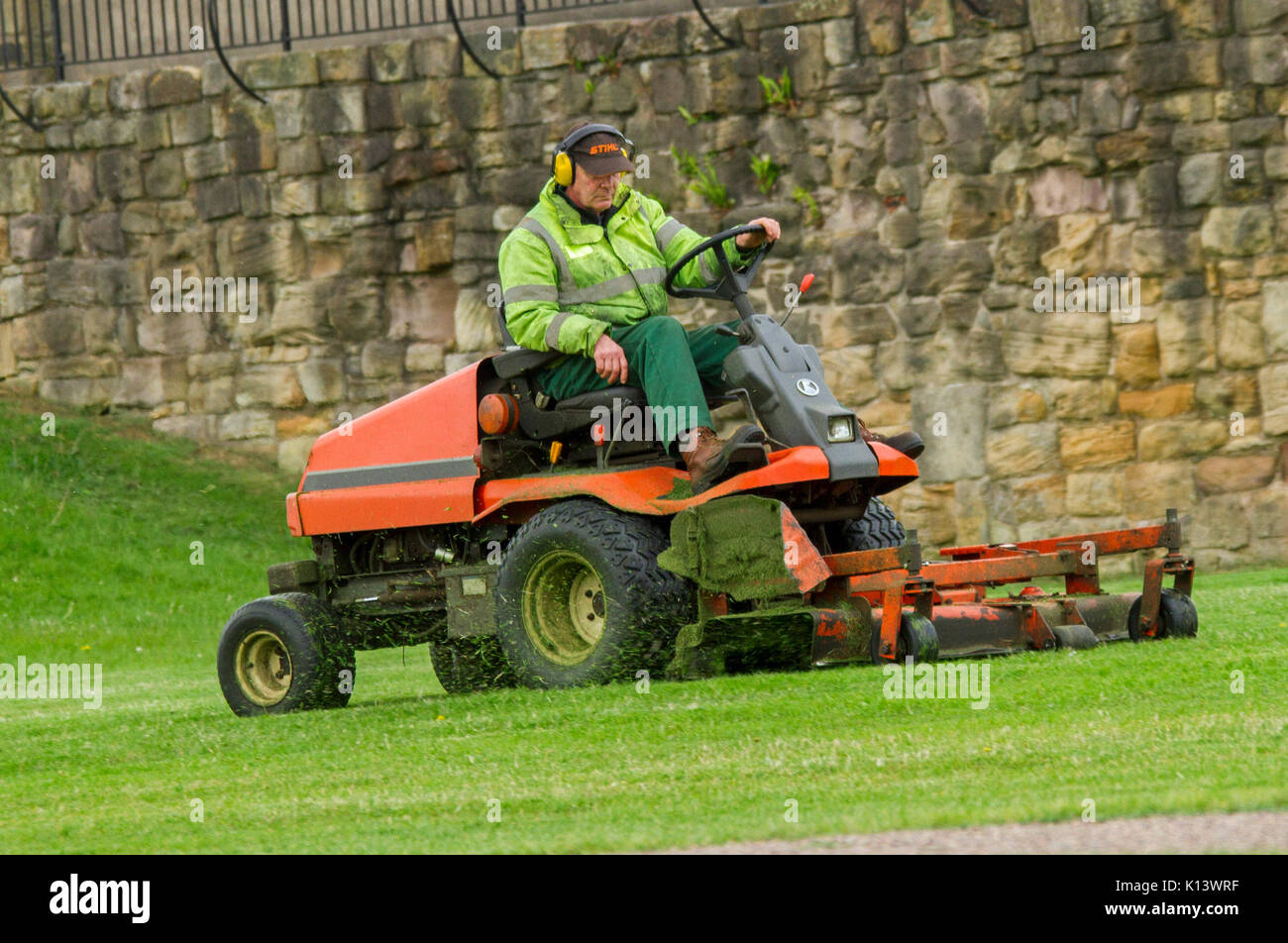 Man wearing high visibility clothing & ear protection using large ride-on mower to cut grass of lawn in public park Stock Photo