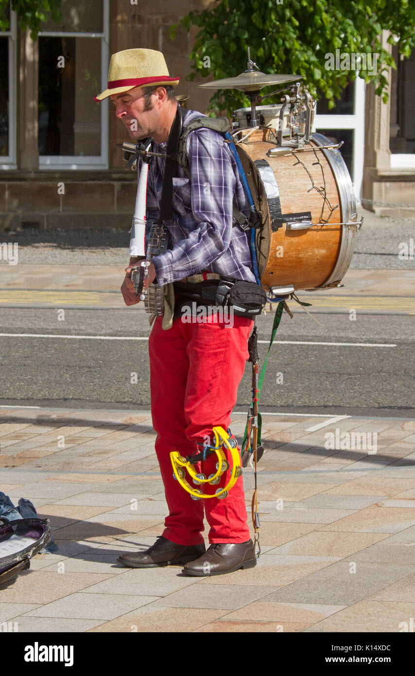 Musician wearing colourful clothing with foot operated drum on back and playing guitar busking in street in Oban, Scotland Stock Photo
