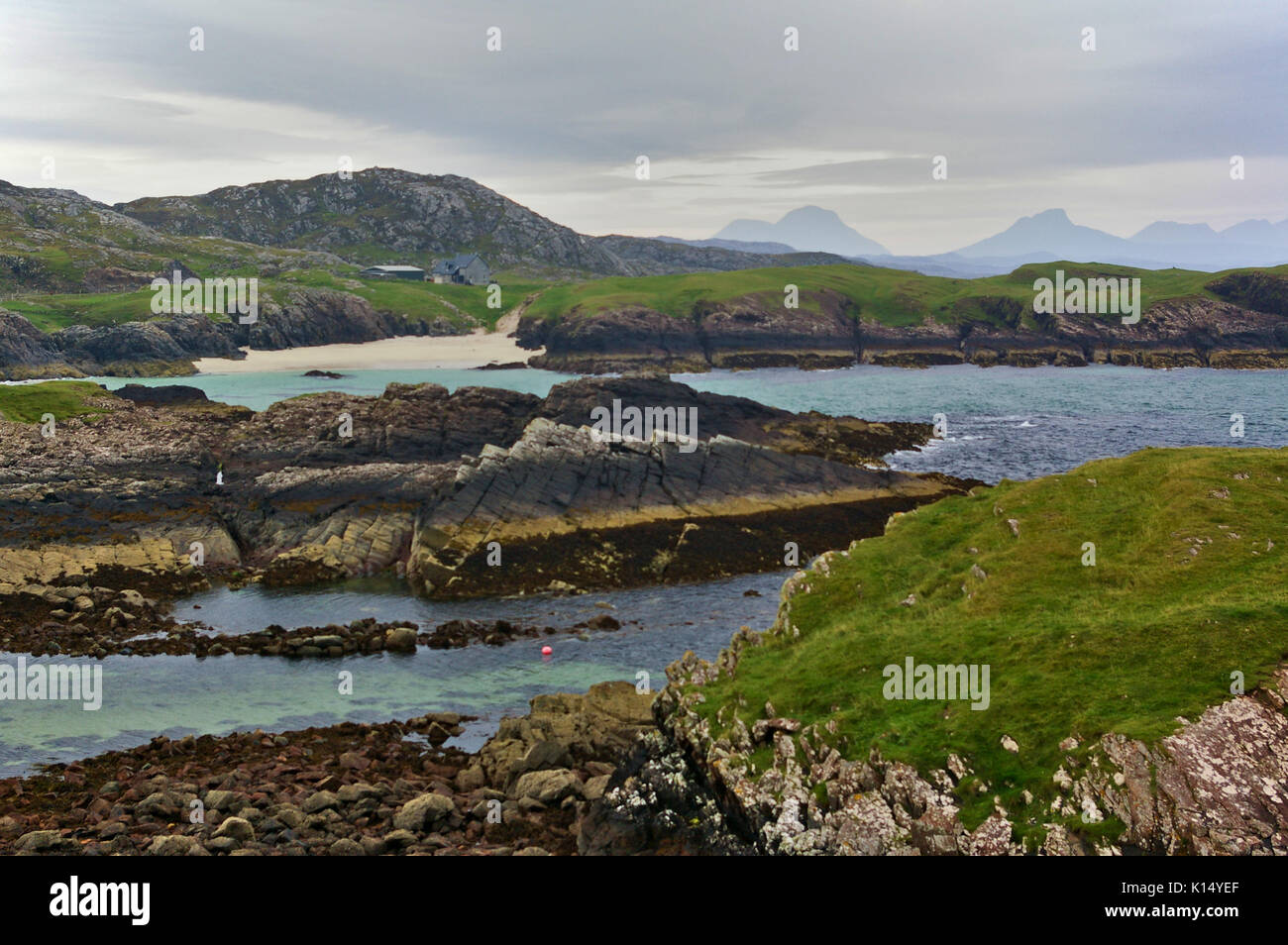 Rocky seashore with patches of green grass and small beach and mountain range in the background near Clachtoll Beach, Sutherland, Scotland Stock Photo