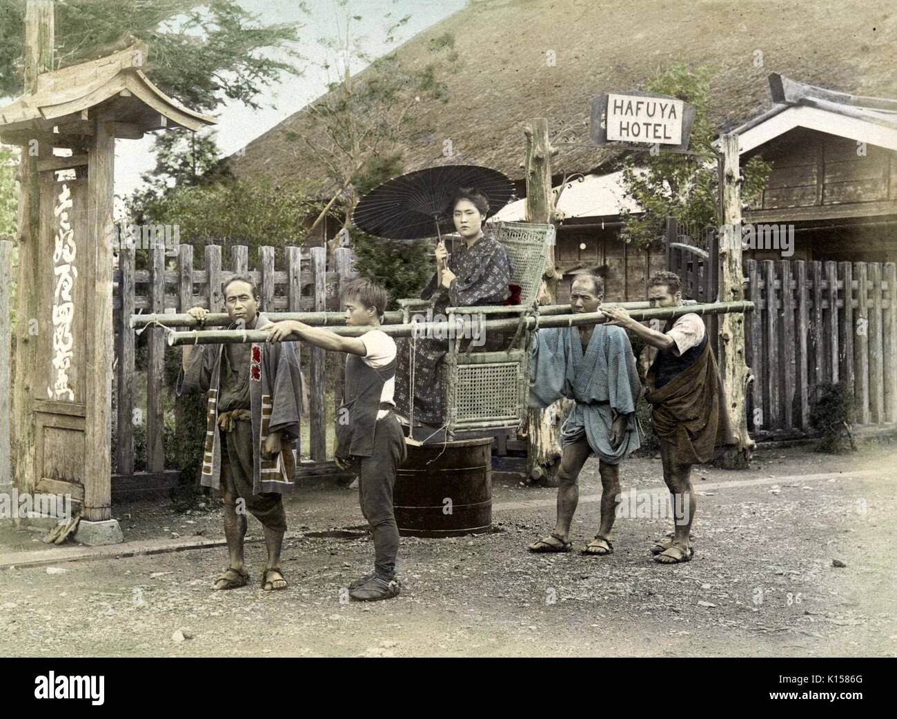 A woman in a fine kimono, holding an umbrella, is being transported in a kago that consists of a chair suspended from two long sections of bamboo, four men are carrying her along a rocky road, the background consists of a fence, hill side, and a building with a sign that reads Hafuya Hotel, Hakone, Japan, 1894. From the New York Public Library. Stock Photo