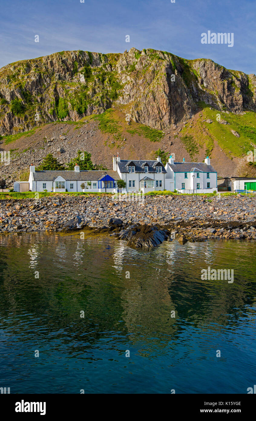 Stunning view of village of Ellenabeich / Easdale, Seil Island, Scotland, with white cottages at base of rocky cliff reflected in calm waters of ocean Stock Photo