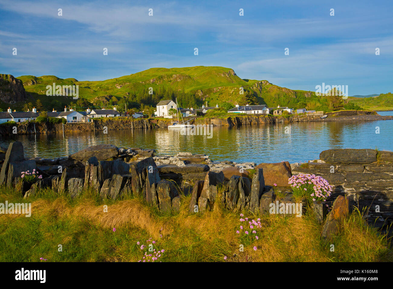 Stunning view of village of Ellenabeich / Easdale, Seil Island, Scotland, with white cottages at base of rocky hill & beside calm blue waters of ocean Stock Photo