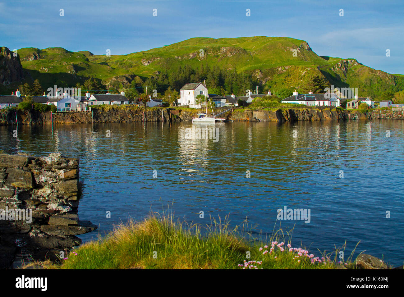 Stunning view of village of Ellenabeich / Easdale, Seil Island, Scotland, with white cottages at base of rocky hill & beside calm blue waters of ocean Stock Photo