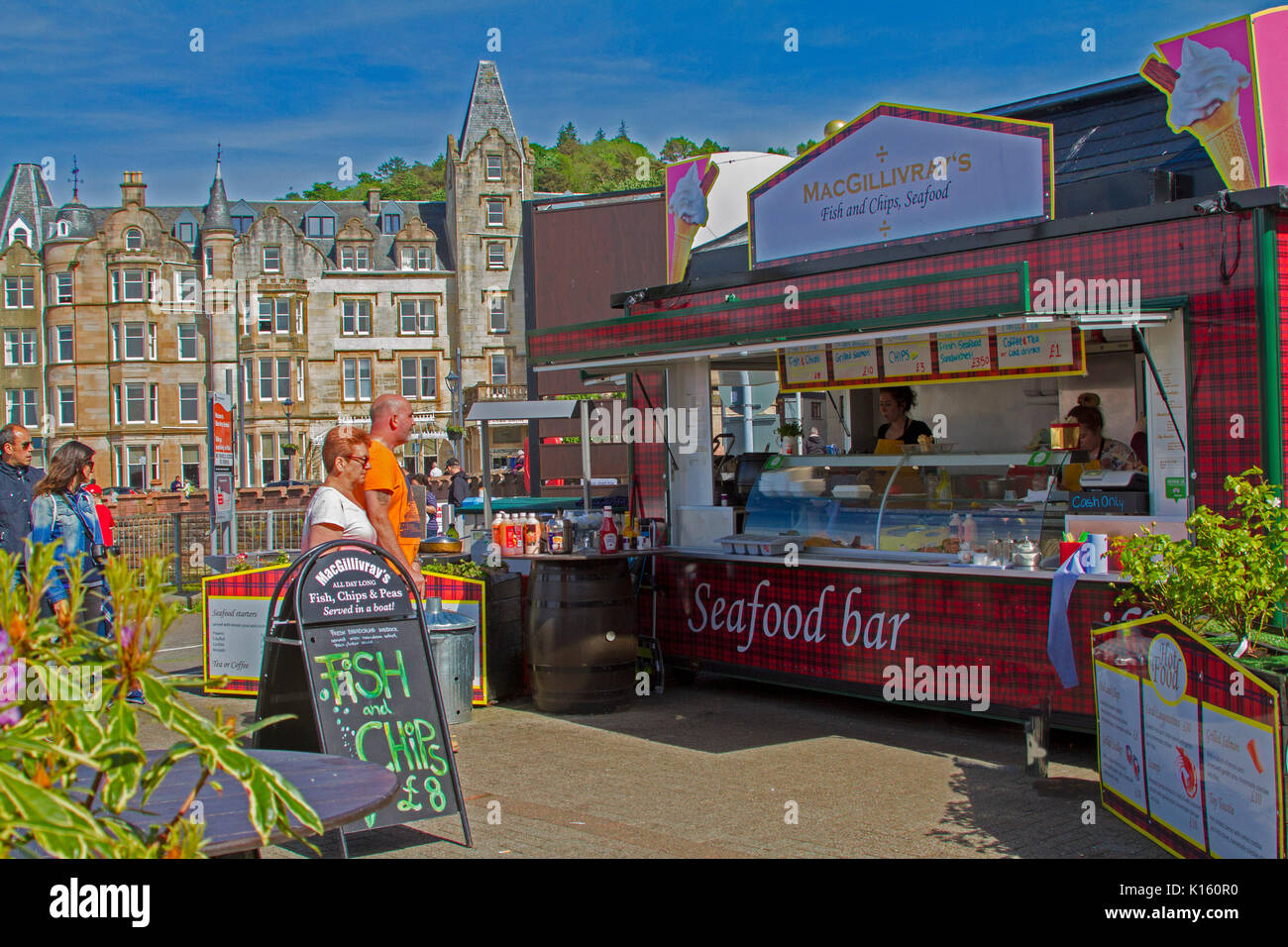 Seafood bar / fish and chip shop beside harbour at Scottish town of Oban, with people looking at signs and buildings of town in background Stock Photo