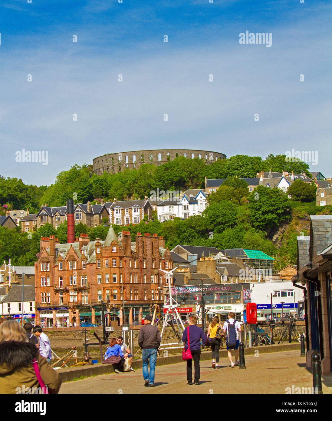 Scottish town of Oban with McCaig's tower on hilltop and people wandering beside harbour and waterfront buildings Stock Photo