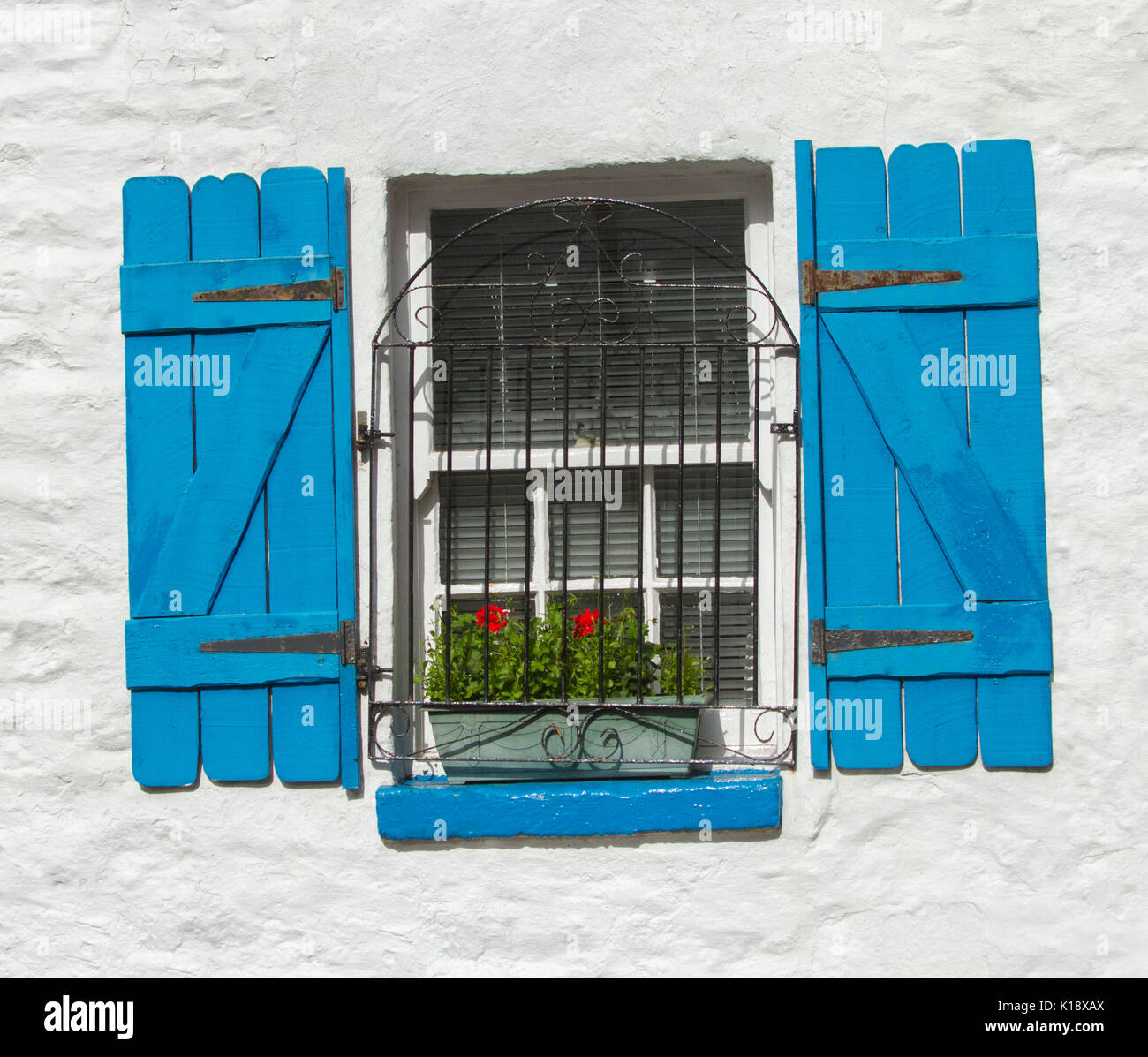 Window with decorative wrought iron security grill, window box with flowers, and bright blue wooden shutters against white painted wall Stock Photo