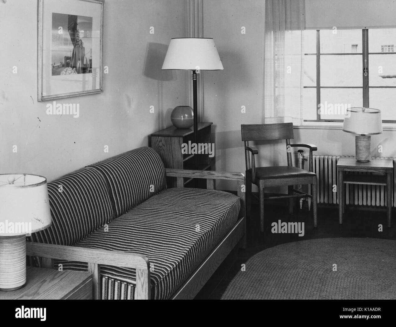 Living room containing a small, striped couch, a wood chair, two wood side tables, two table lamps, a floor lamp, and an oval rug, Greenbelt, Maryland, 1936. From the New York Public Library. Stock Photo