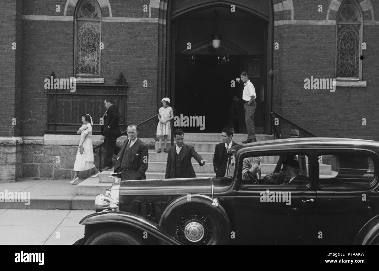Small gathering of people outside of a church, car with two men parked in front, men in three piece suits next to the car, a girl dressed in white wearing a hat on the church steps, New Hampshire, 1937. From the New York Public Library. Stock Photo