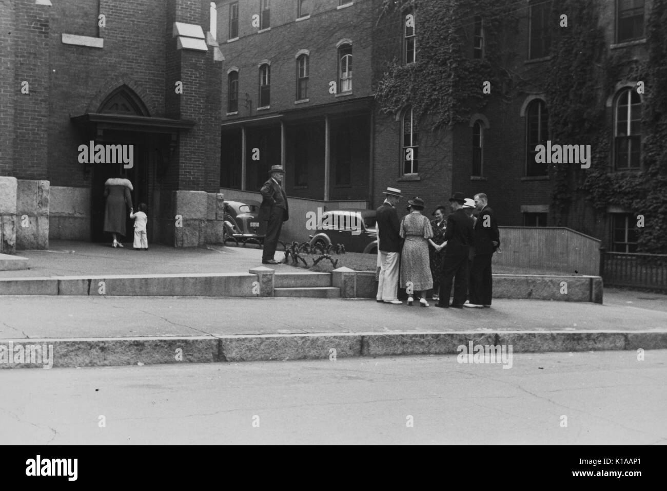 A gathering of adult men and women in front of a church on a Sunday, men in hats and suits and women in hats and long dresses, a mother and child, holding hands, entering the church, Manchester, New Hampshire, 1937. From the New York Public Library. Stock Photo