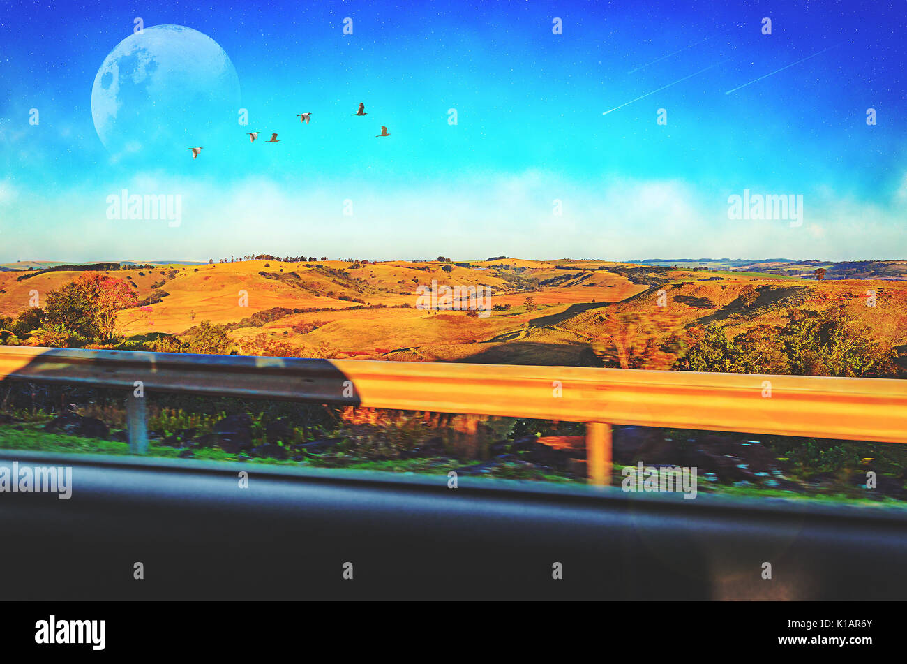 View from the car window of a landscape with farm fields, hills, a deep blue sky, some clouds, a big moon, birds flying, stars on sky and shooting sta Stock Photo