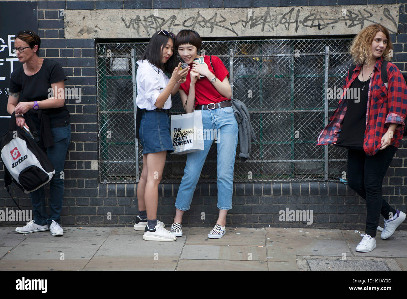 LONDON, ENGLAND - August 12, 2017 Two smiling girls are examining photos in the phone near the brick wall at Bricklane Stock Photo