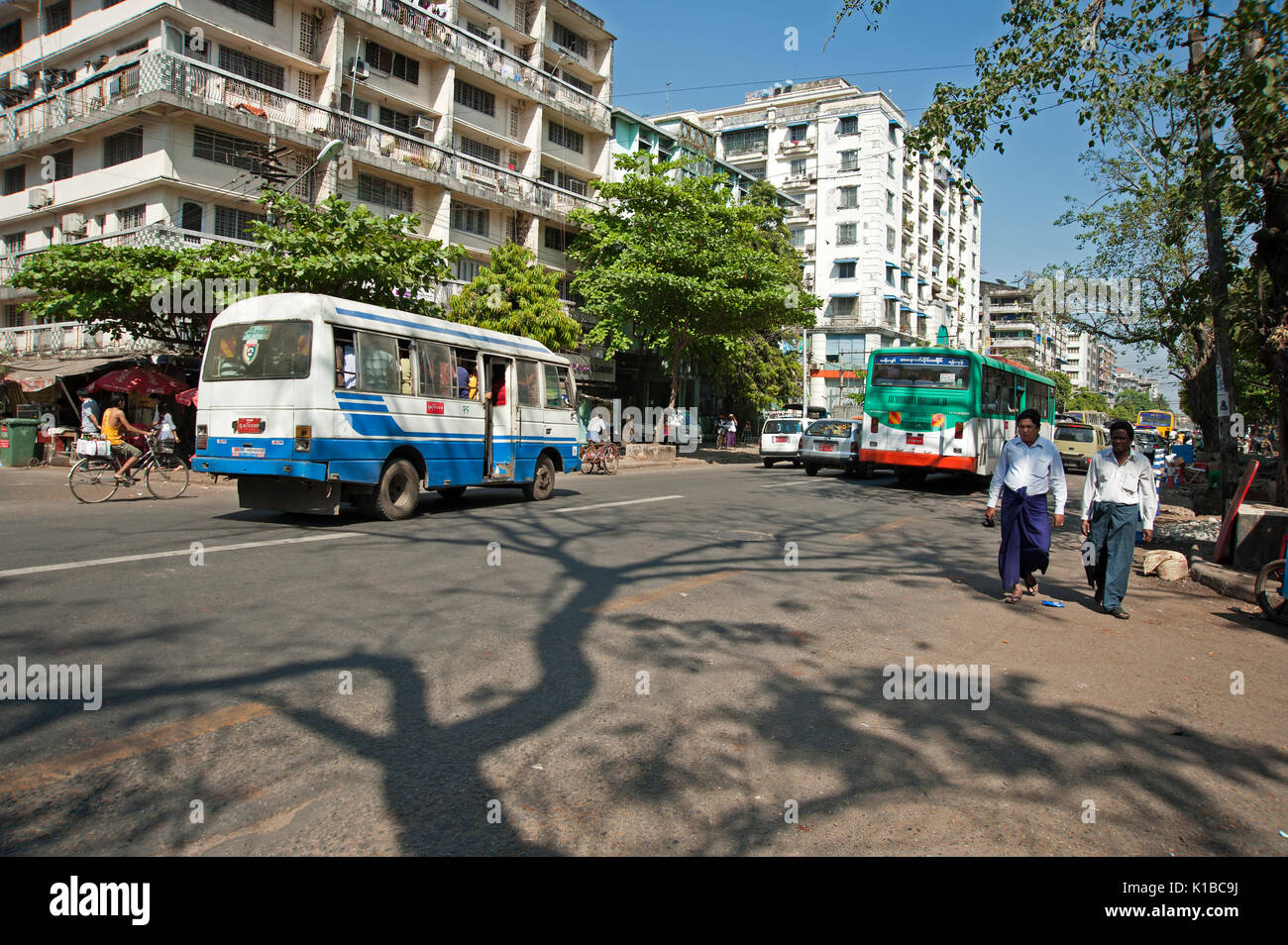 Two Burmese men walk along a busy road in Yangon Myanmar with modern buildings and buses behind them Stock Photo